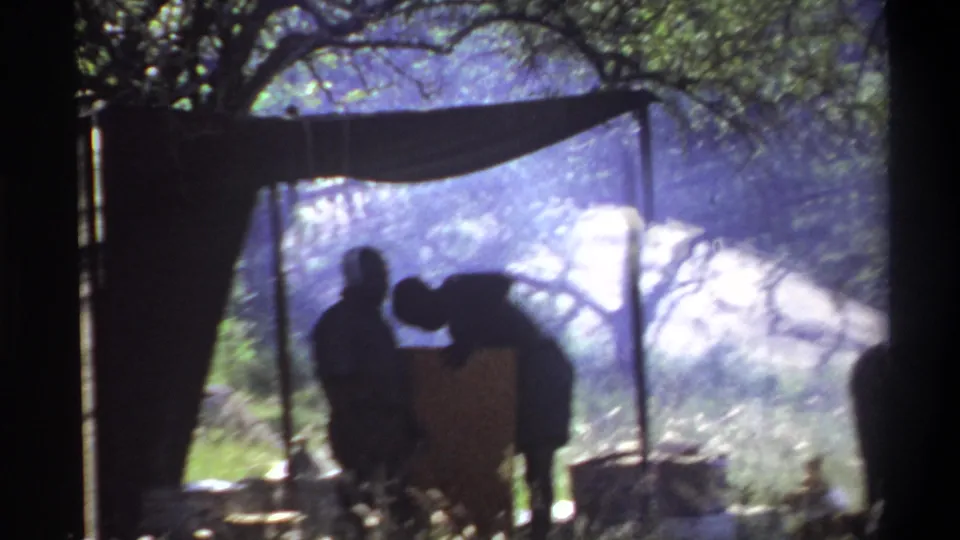 1969 Lagos 8mm frame of silhouetted campsite activity with two people, a canopy, cooking gear, smoke, grass, and trees.