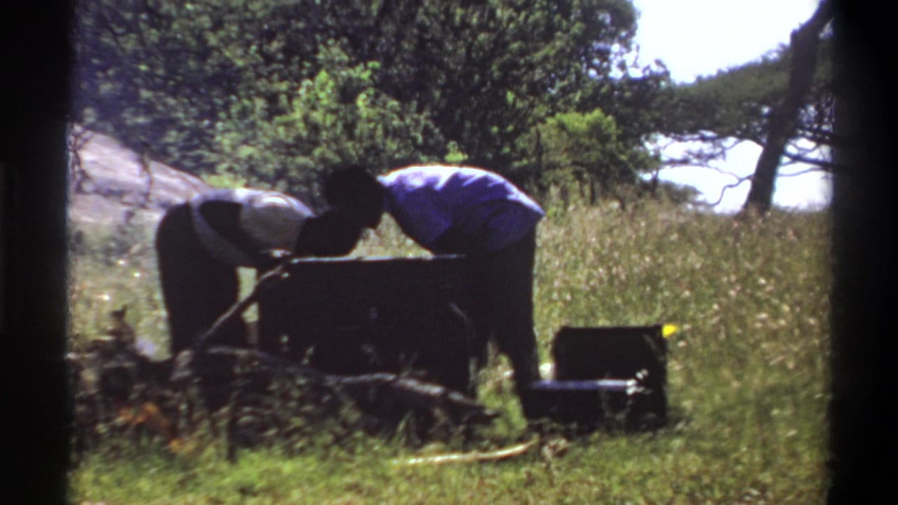 1969 Lagos frame with two silhouetted figures leaning over a large container beside camp cases and tall grass.