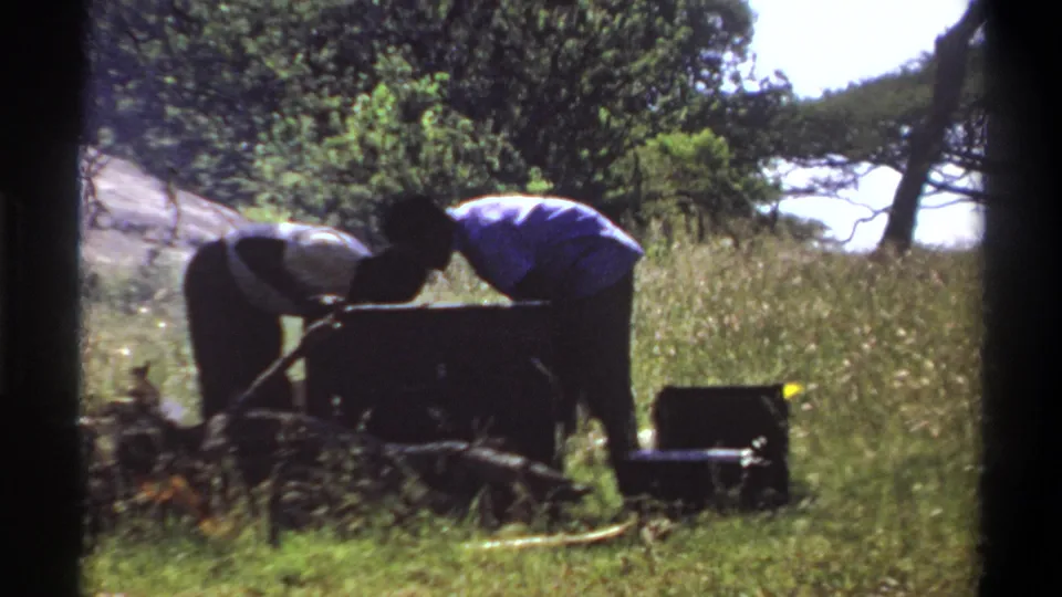 1969 Lagos frame with two silhouetted figures leaning over a large container beside camp cases and tall grass.
