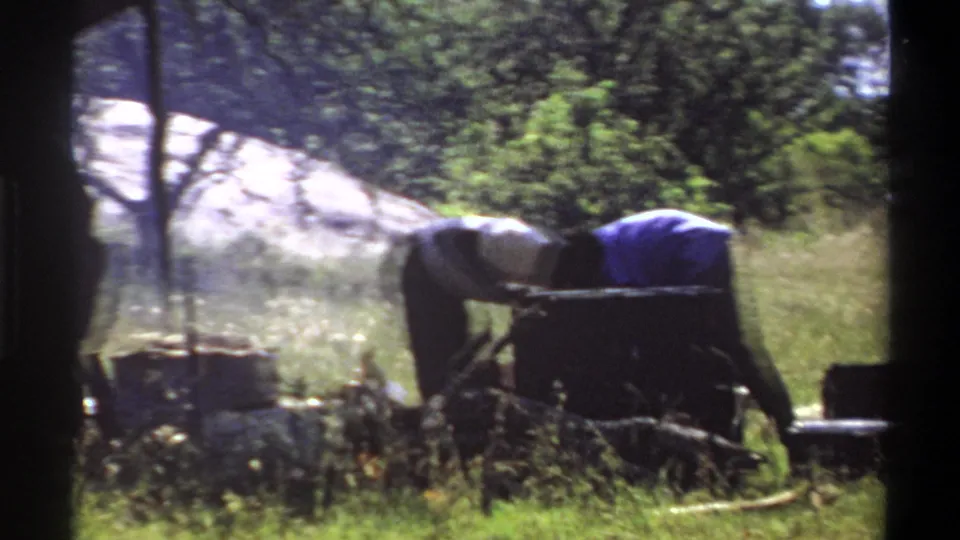 1969 Lagos home-movie frame showing people bent over campsite gear, with a canopy edge, smoke or haze, grass, and trees.