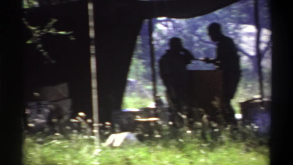 1969 Lagos frame showing dark silhouettes beside a large camp container under a canopy, with cases, grass, and wooded background.