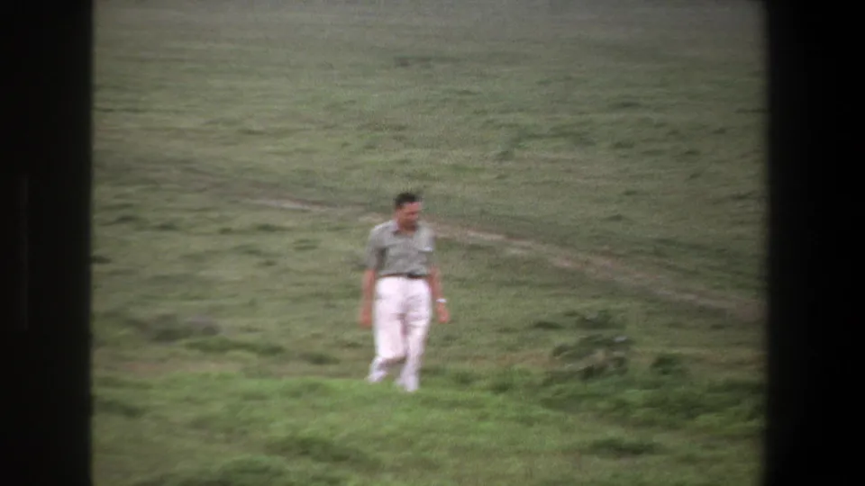 1969 Pretoria home-movie frame showing a man in a pale shirt and white pants walking across uneven grassland.