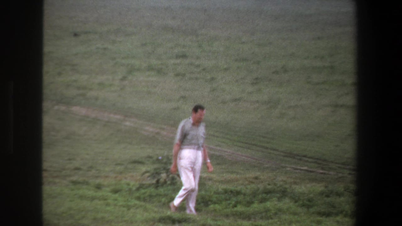 Man in white trousers walking across a grassy field near a dirt track.