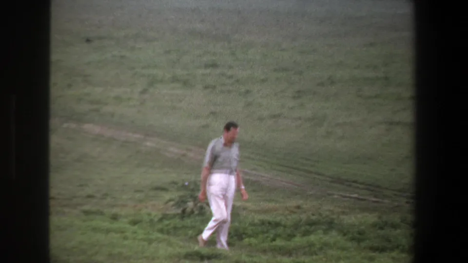 In 1969 Pretoria home-movie footage, a man in white trousers walks through open grassland with a faint dirt track behind him.
