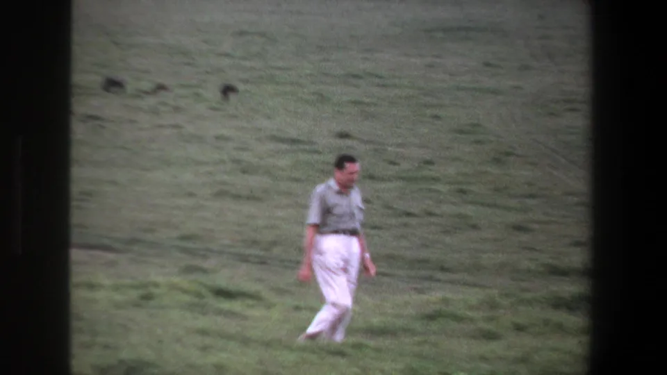 1969 Pretoria home-movie frame showing the man moving through uneven grass with distant animals softened by film grain.