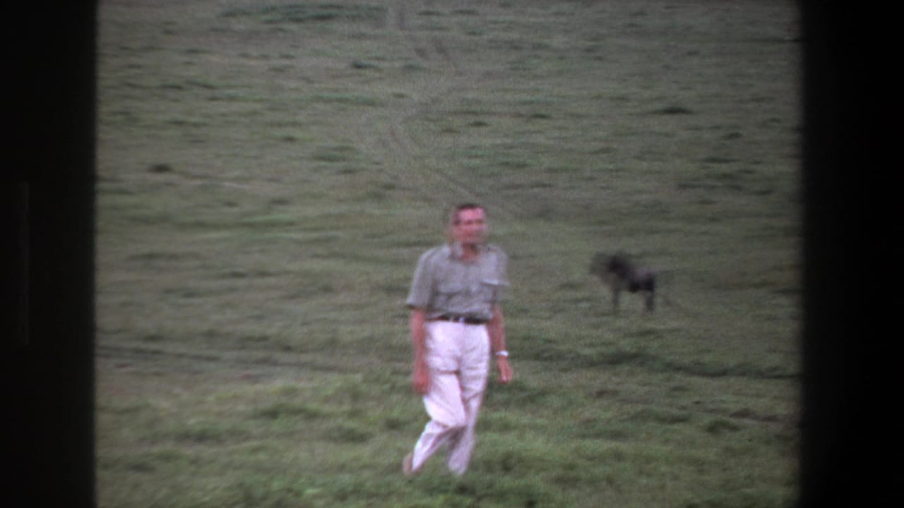 Man in white trousers walks across a grassy field with a dark animal behind him.