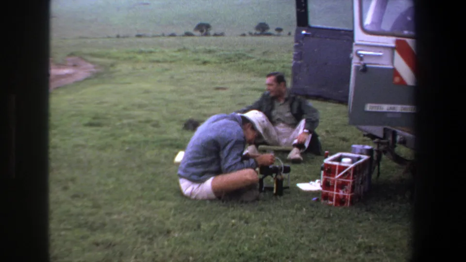 Two men sit near camera gear on a grassy area beside a parked vehicle, with a red cooler in the foreground.