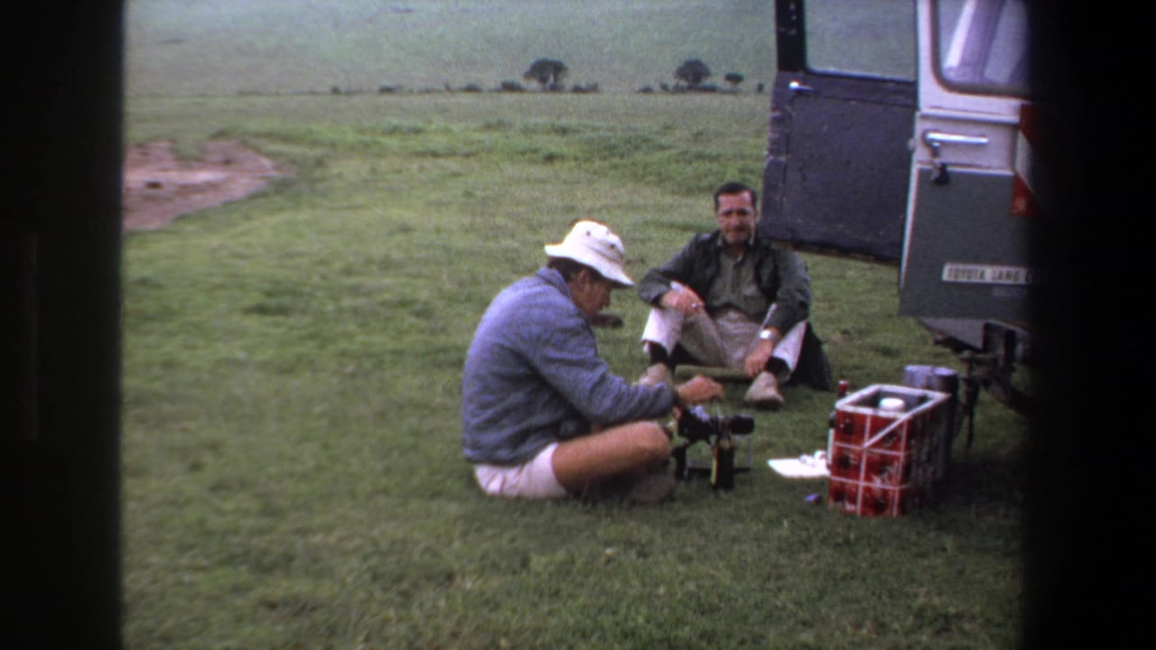 In 1969 Pretoria-area home-movie footage, two adult men pause beside an open vehicle with a red cooler and small equipment on the grass.