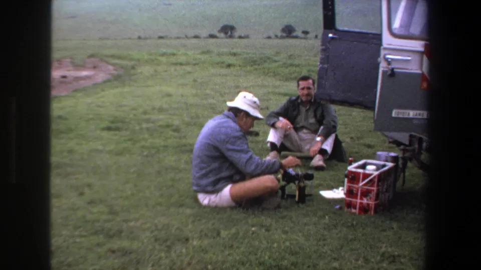 In 1969 Pretoria-area home-movie footage, two adult men pause beside an open vehicle with a red cooler and small equipment on the grass.