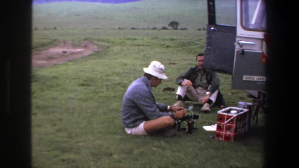 The two adult men continue a quiet camping stop on grass beside the open vehicle, with a red cooler crate and supplies visible.