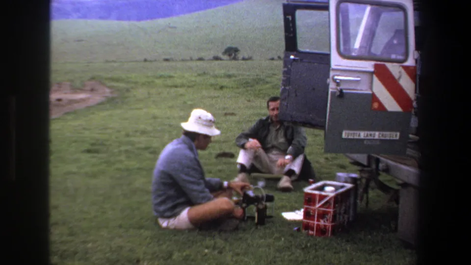 The rear of the open vehicle is more prominent as two men sit nearby with camping supplies on a grassy Pretoria-area stop in 1969.