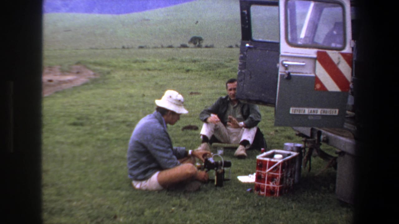 1969 Pretoria home-movie frame showing two adult men preparing camera equipment beside an open silver Land Cruiser in grassy hills.