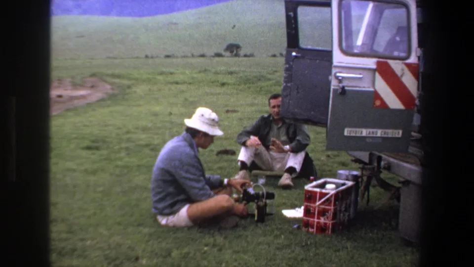 1969 Pretoria home-movie frame showing two adult men preparing camera equipment beside an open silver Land Cruiser in grassy hills.