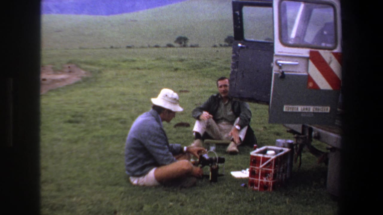 Man in a white hat handles camera equipment while another man sits near the open rear of a vehicle.