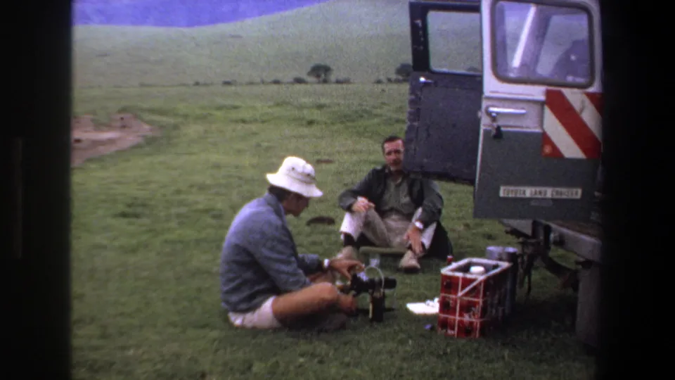 Two adult men in a grassy Pretoria setting, one adjusting ground-level camera gear near supplies and an open Land Cruiser.