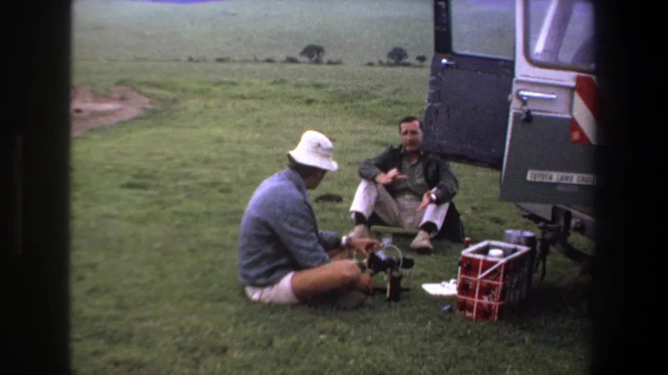 1969 South Africa frame with two adult men and visible safari supplies beside a silver Land Cruiser in open grassland.