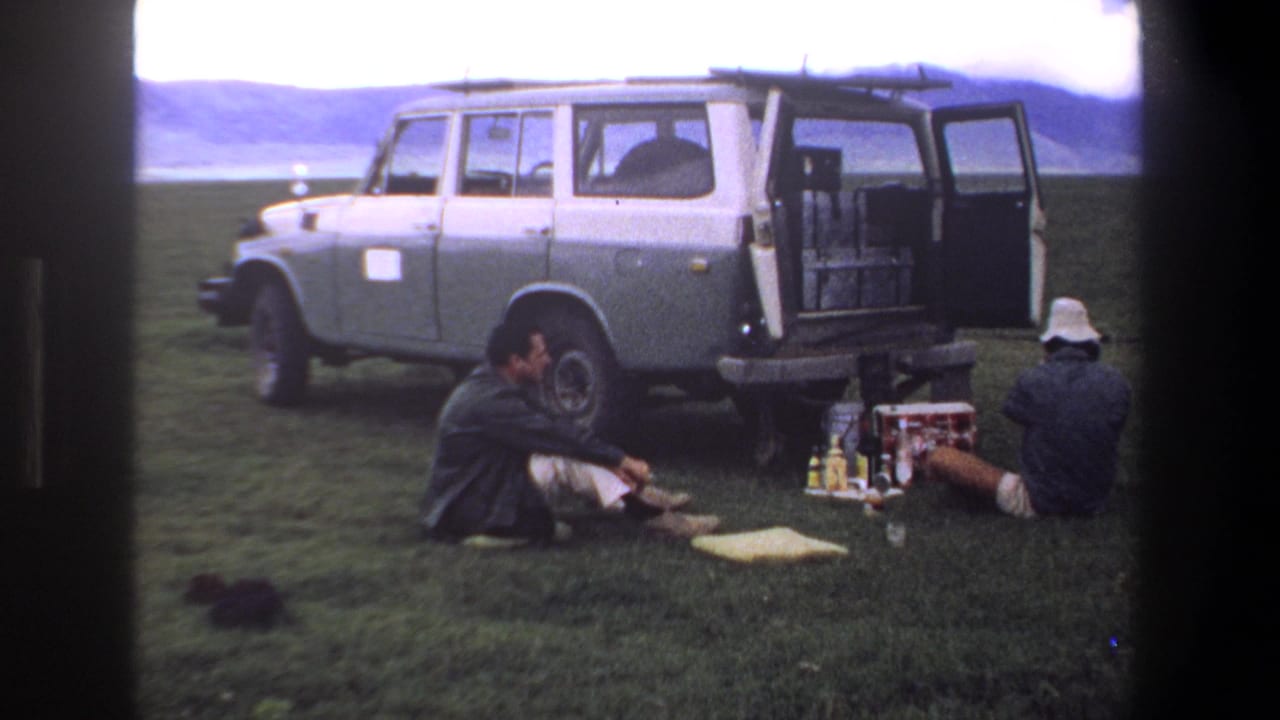 1969 Pretoria home-movie frame showing two adult men seated on grass near picnic supplies beside a parked two-tone station wagon.