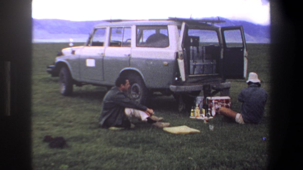 Two seated men and picnic supplies beside an open station wagon on grass.