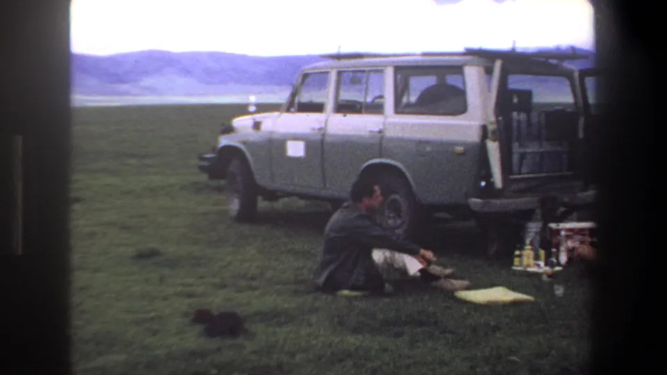 1969 Pretoria home-movie frame with one seated man visible beside picnic supplies and a two-tone station wagon in open grassland.