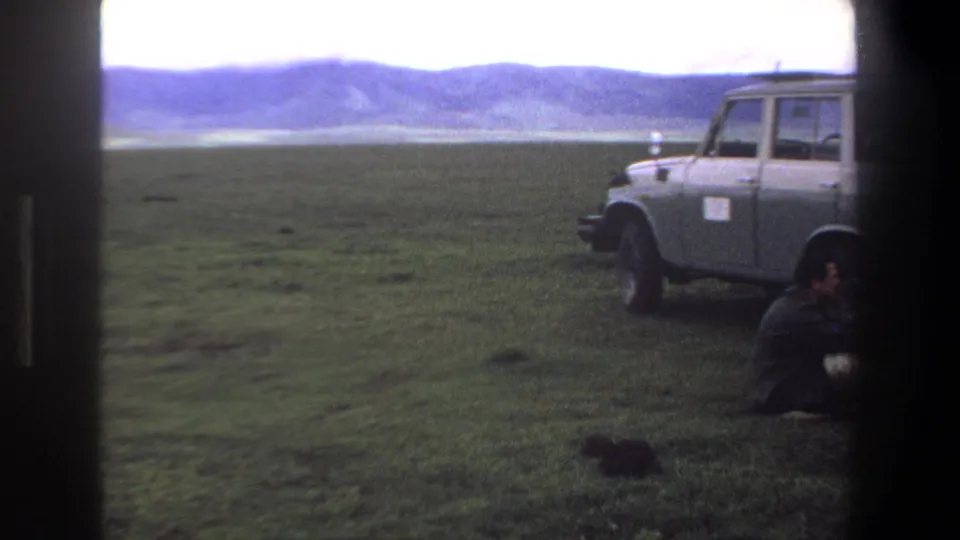 1969 Pretoria frame with the vehicle partly cropped at right and one seated man on grass against distant hills.