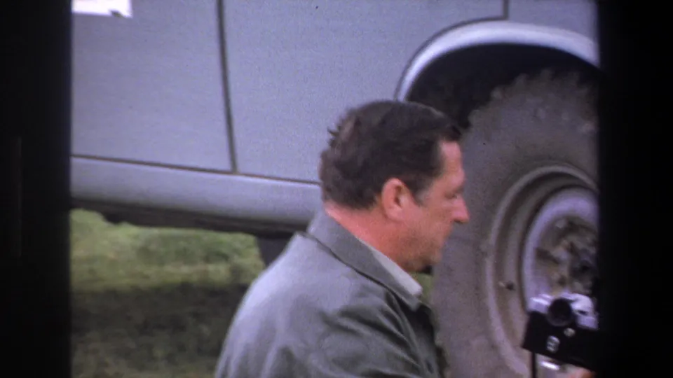 An adult man stands beside a light-colored utility vehicle, with a small camera-like object visible near the wheel area.