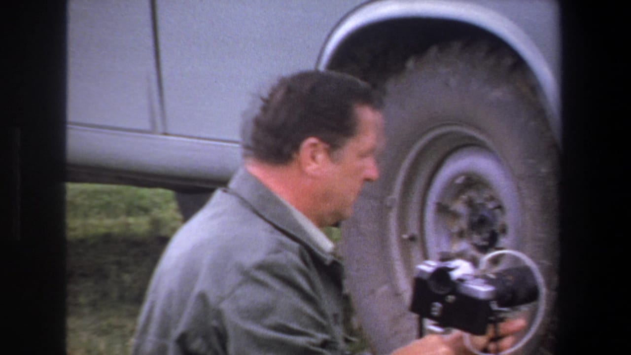 Man in a light jacket crouches beside a truck wheel while holding a small object.