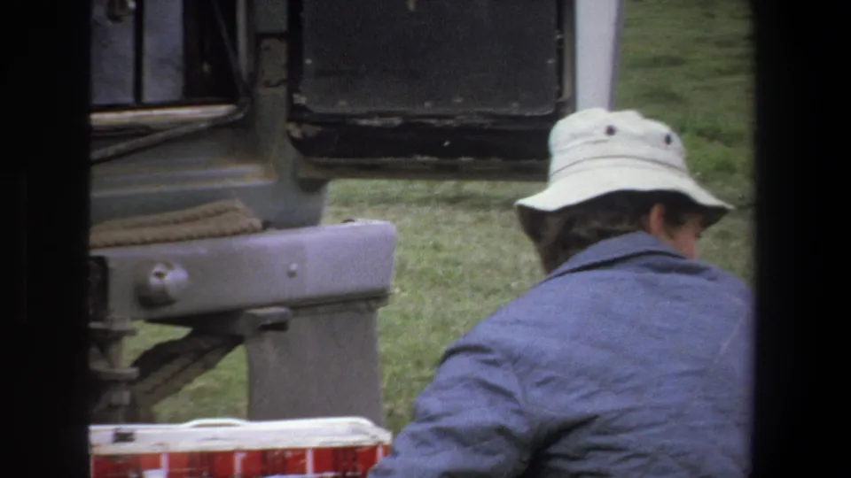 An adult man wearing a pale bucket hat bends beside the rear of the truck during the 1969 Pretoria roadside repair scene.