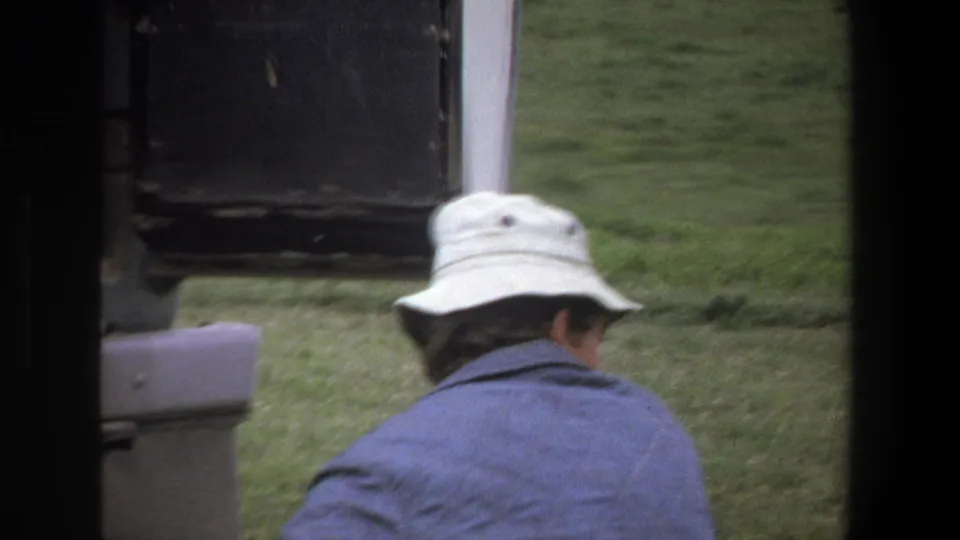 In 1969 Pretoria safari footage, a person in a white bucket hat and blue jacket looks out from a vehicle toward grassy terrain.