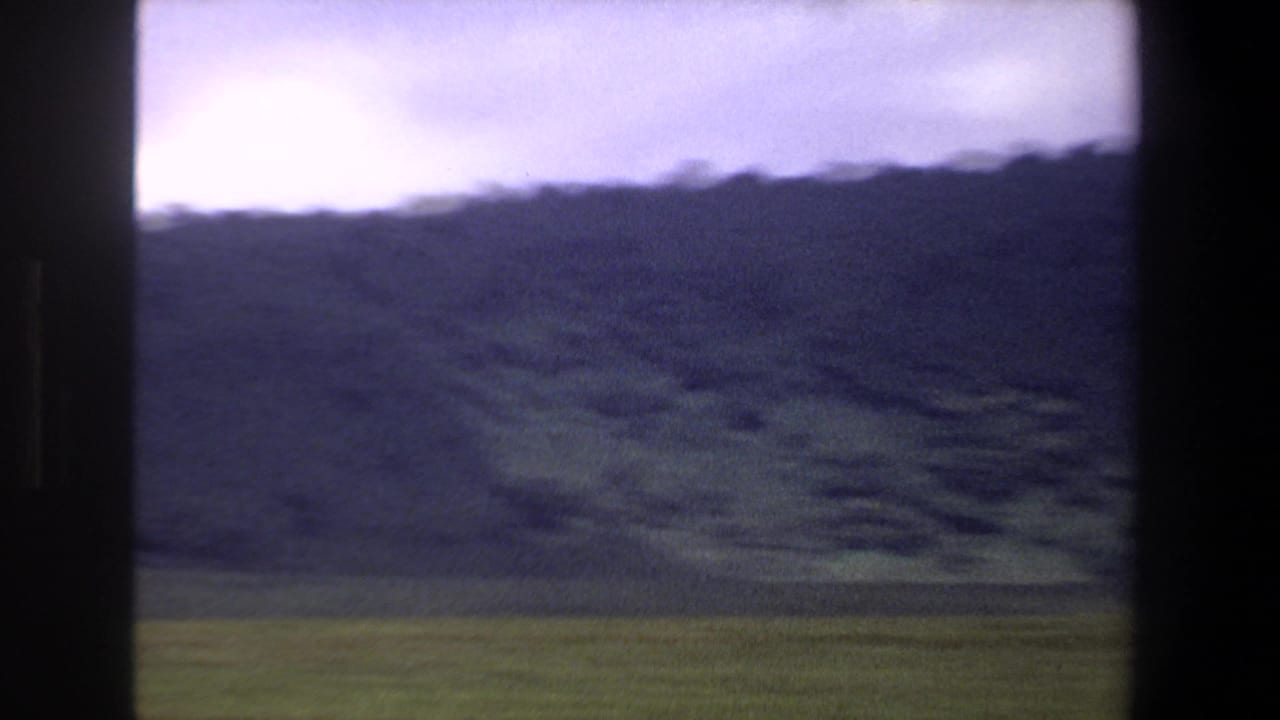 1969 Pretoria home-movie frame showing a green field and dark mountain slope beneath an overcast sky.