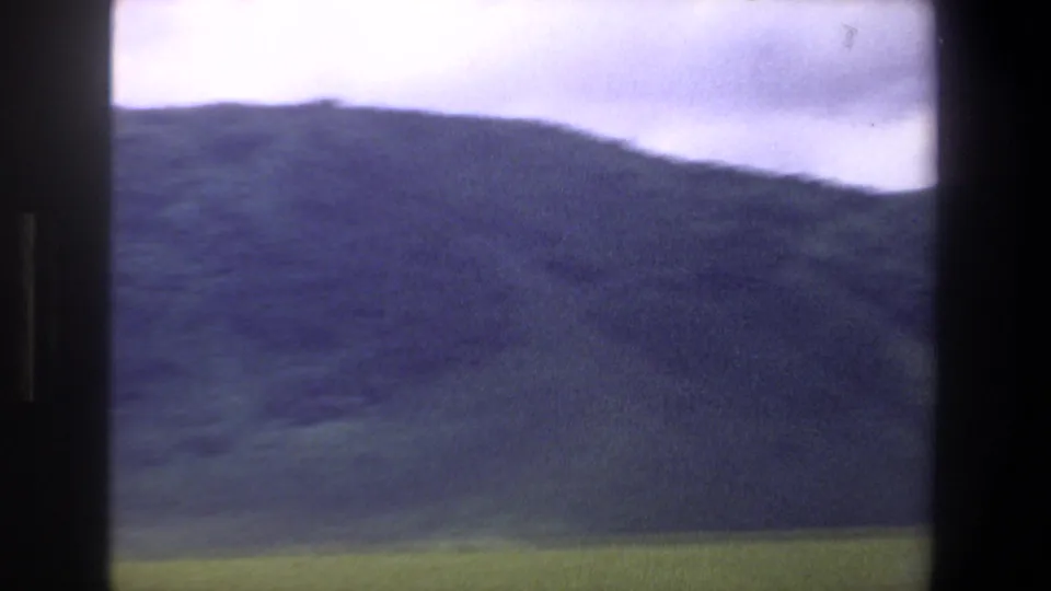 1969 South Africa home-movie still showing a blurred dark slope, green foreground, and pale clouded sky.
