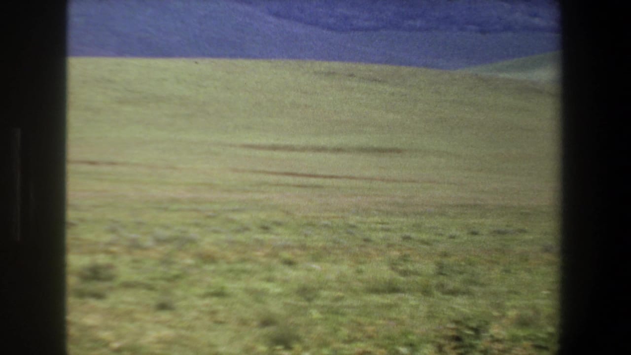 1969 Pretoria home-movie frame showing open green meadow, low dark hills, and a blue-purple sky through dark window or film edges.