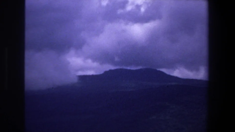 1969 South Africa home-movie landscape frame showing distant hills under heavy clouds beside a blue-toned body of water.