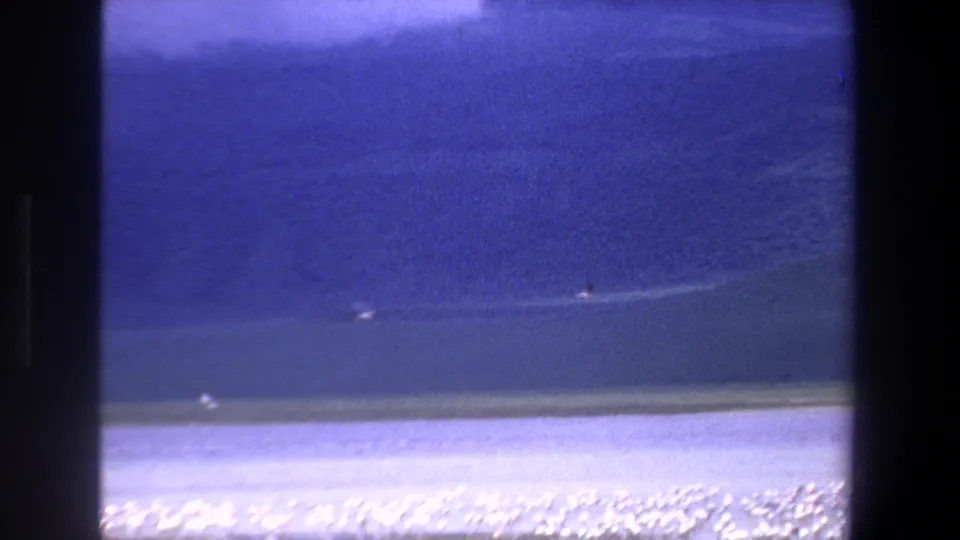 1969 South Africa 8mm frame showing a blue body of water, pale shoreline, and small birds scattered across the surface.