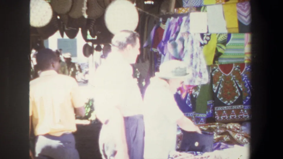 1969 Pretoria home-movie frame of shoppers at an outdoor market stall with hanging baskets, bright cloth, and patterned textiles.