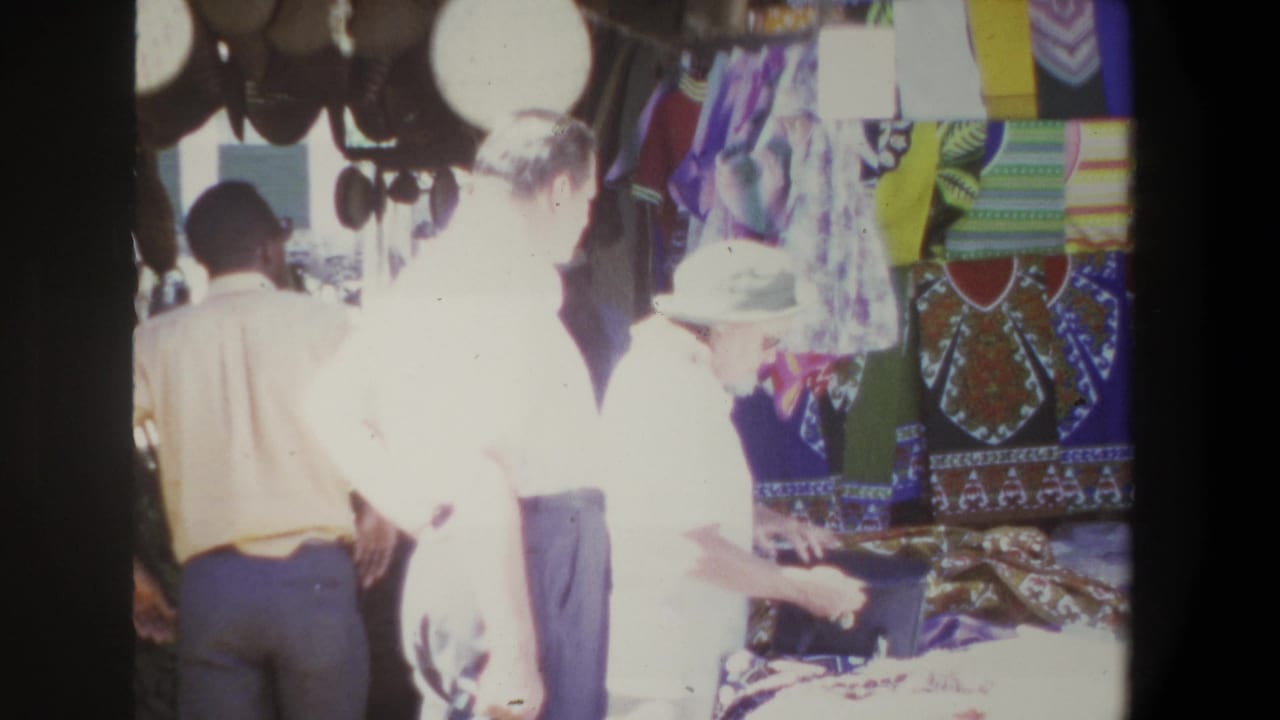 In Pretoria, South Africa, 1969, adults stand at a bazaar stall with hanging hats, patterned fabrics, and bright overexposed daylight.