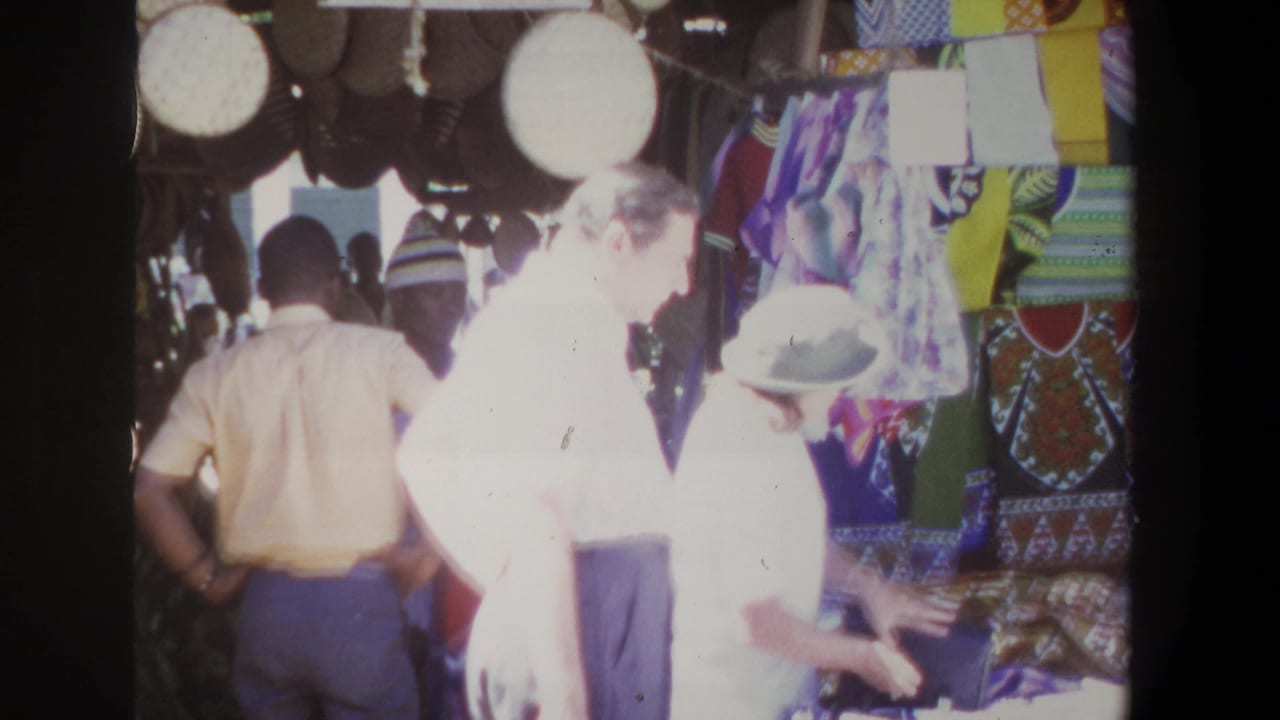 A man and woman stand near patterned fabrics and goods at a market stall.