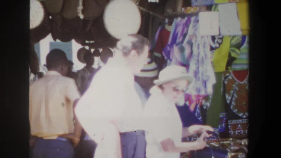 The handheld 8mm view shows adults browsing a Pretoria market display with patterned fabric panels, hats, and goods on a table.