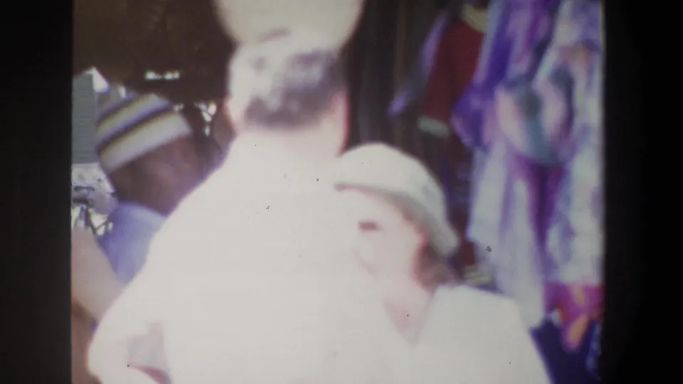 A soft, close 1969 Pretoria bazaar frame catches adults moving near the camera, with colorful textiles and shaded stall details behind them.