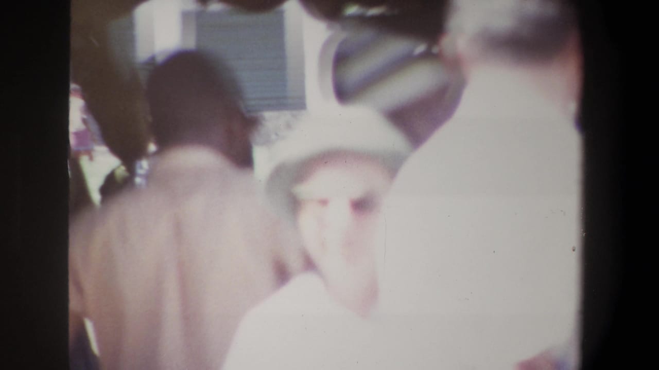 1969 Pretoria home-movie frame showing crowded market browsing, with a light-hatted figure near the lens and other shoppers partly silhouetted.