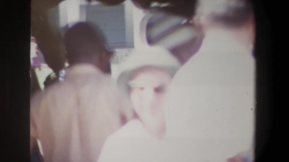 1969 Pretoria home-movie frame showing crowded market browsing, with a light-hatted figure near the lens and other shoppers partly silhouetted.
