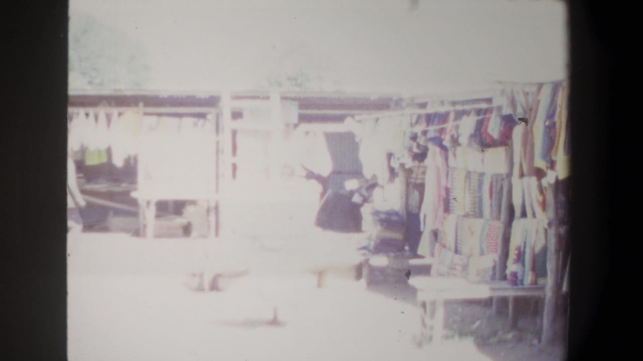 1969 Pretoria market view with hanging garments, shaded stalls, wooden structures, and overexposed shoppers moving through the aisle.