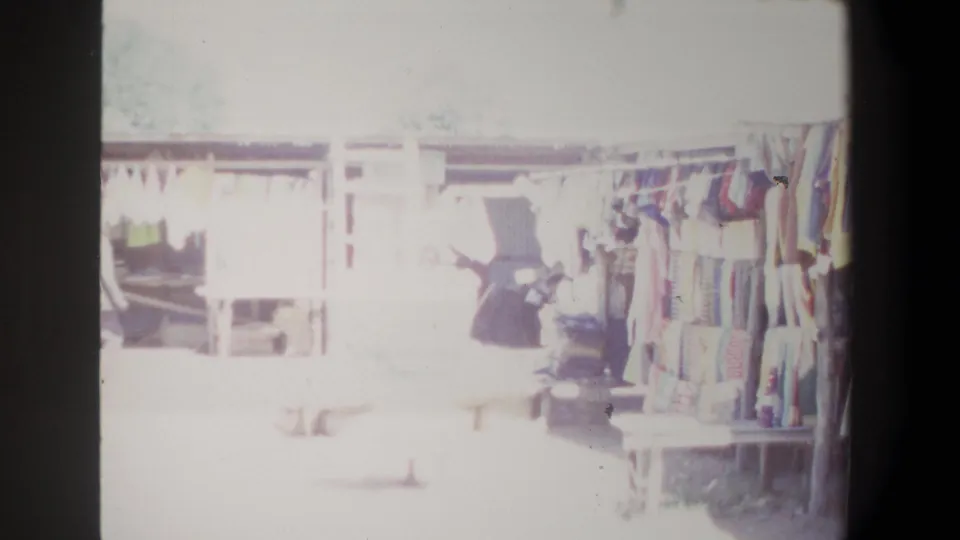 1969 Pretoria market view with hanging garments, shaded stalls, wooden structures, and overexposed shoppers moving through the aisle.