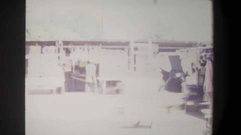1969 Pretoria home-movie frame of a washed-out market aisle, showing wooden stall fronts, textiles, and figures partly lost in bright sunlight.