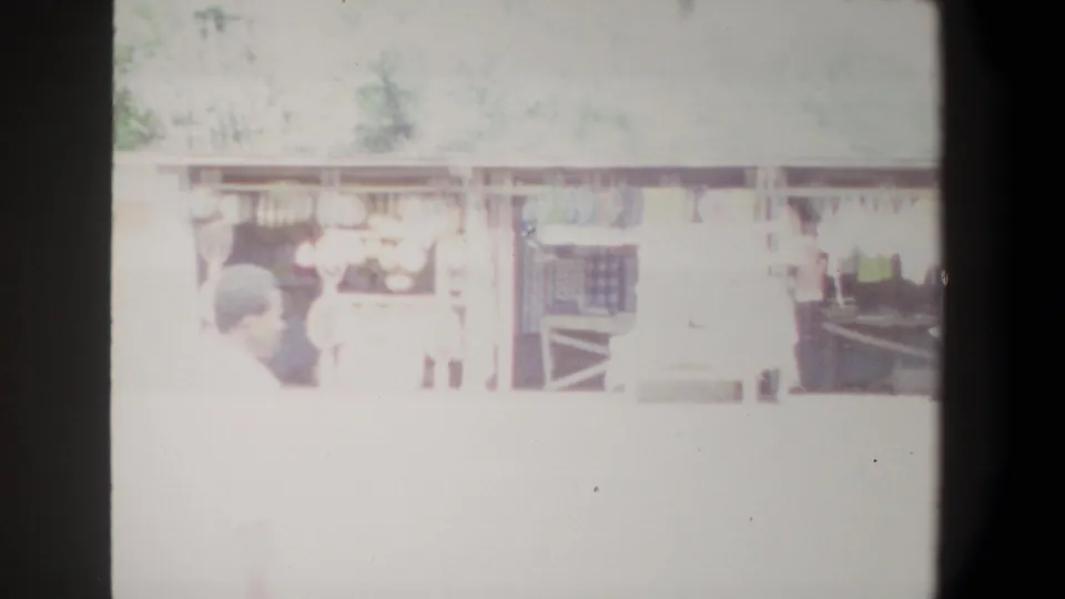 1969 Pretoria home-movie frame showing pedestrians near a long open-sided building, partly obscured by exposure bloom and film borders.