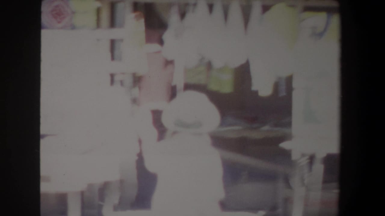 1969 Pretoria home-movie frame showing a bright outdoor market scene with a hat-wearing person near textile displays.