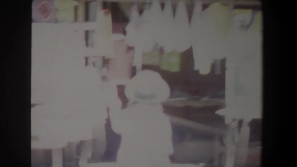 1969 Pretoria home-movie frame showing a bright outdoor market scene with a hat-wearing person near textile displays.