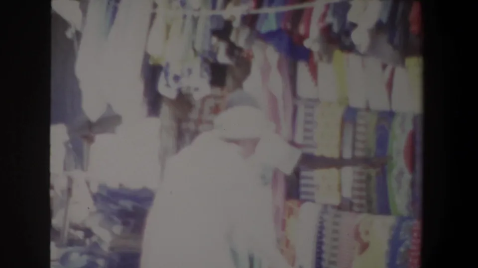 A 1969 Pretoria home-movie frame shows a hat-wearing shopper examining colorful textiles at an outdoor market stand.