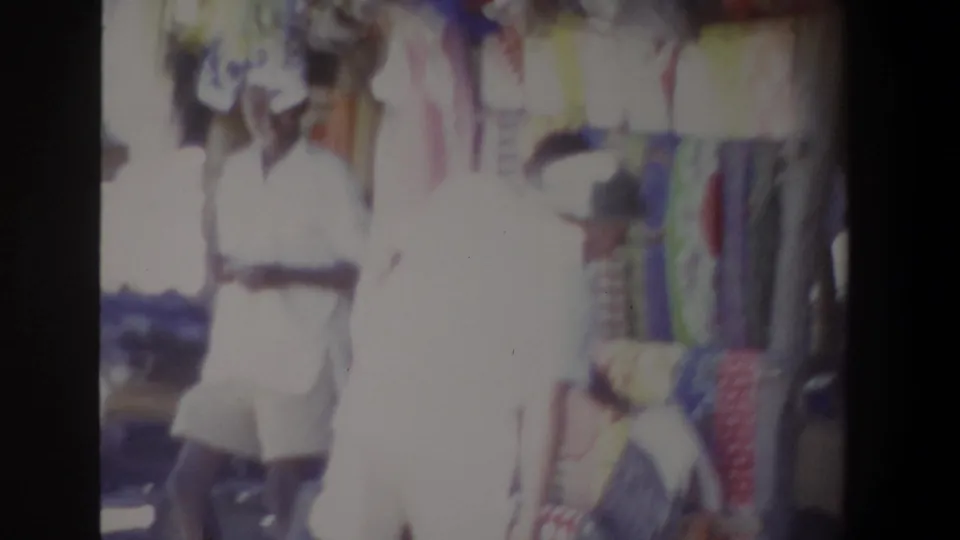 In the supplied 1969 Pretoria market footage, a hat-wearing person stands before a display of colorful hanging textiles.