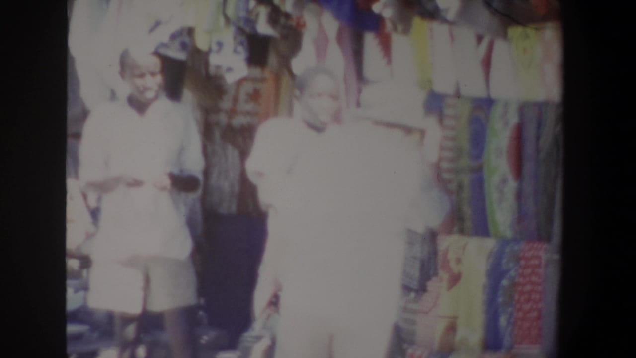 In 1969 Pretoria market footage, two figures appear beside a colorful clothing stall, with dresses and cloth hanging behind them.