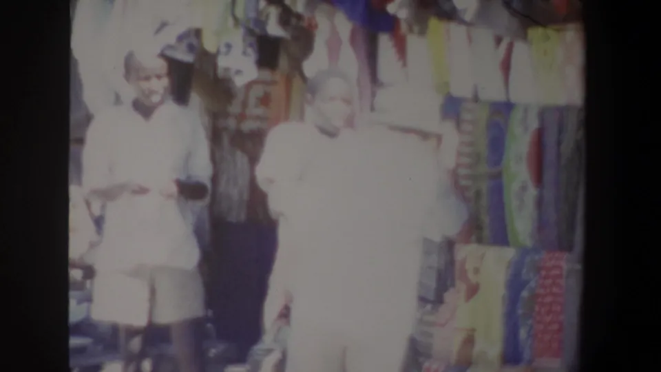 In 1969 Pretoria market footage, two figures appear beside a colorful clothing stall, with dresses and cloth hanging behind them.